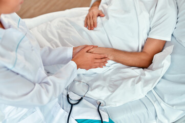 A nurse sits near a mature male patient and holds his hand to show her care and support in a modern hospital ward.