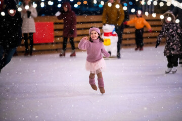 little girl in a pink sweater and skirt is skating on a winter evening on an outdoor ice rink lit...