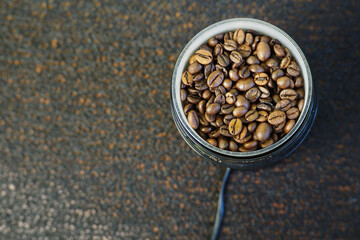 brown whole grains of roasted coffee are poured into a plastic electric coffee grinder on a black textured background top view