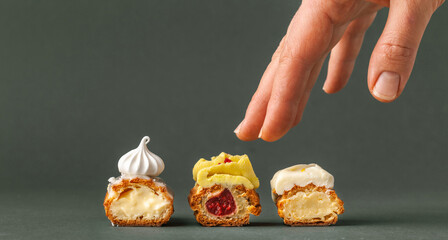 Set of fresh eclairs cut with  butter cream filling and meringue cookies decoration. French pastry with woman hand reaching the cookie on the dark green or grey background.