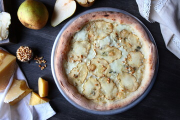 Pizza with dried pears and pine nuts. Traditional Italian pizza. Metal plate for serving pizza. A piece of parmesan and a pear on a dark background. White cotton napkin. The view from the top.