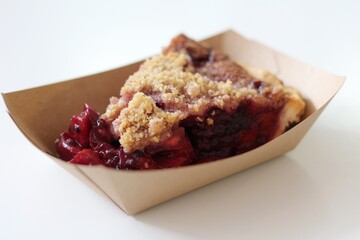 Slice of Red Berry Pie with Crumble Top in Brown Cardboard Boat on White Table