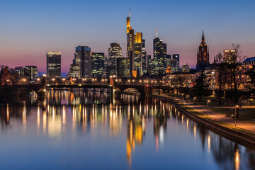 Frankfurt skyline in the evening at the blue hour. River Main with reflections of high-rise buildings. Illuminated commercial buildings with a bridge and park on the river bank