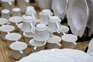 A set of plates and cups in white tone stands on a light wooden table. A stack of ceramic plates, glasses, mugs. Simple design, close-up top view. Place for your text. empty mugs of tea and saucers.