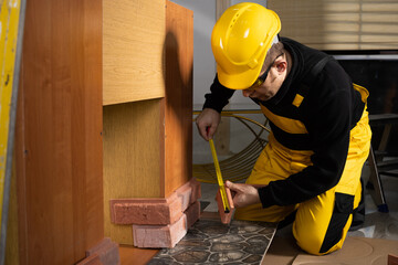 A construction worker in working clothes, kneeling, measures the retractable decorative tiles in the shape of old red brick with a tape measure.