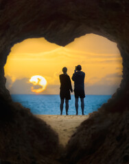 Paar Silhouette schaut am Meer vor einer Herz- förmigen Höhle in den Sonnenaufgang 