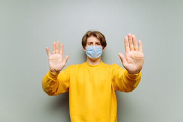 Young man in yellow sweatshirt and medical protective mask shows hands stop gesture on gray wall background. Focus on the palms. guy in a medical mask. Isolated.