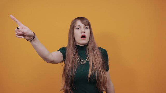Pointing with hand aside and staring. Shocked facial expression. Young attractive woman, dressed green shirt blonde hair, yellow background