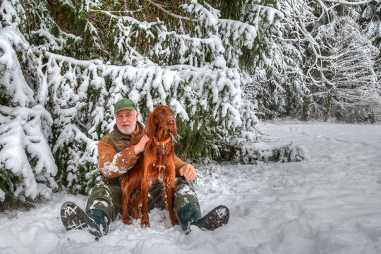 In the snow-covered Black Forest, a hunter sits with his beautiful Irish Setter hunting dog at the edge of the forest in the snow.