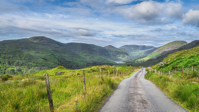 Long country road leading to majestic Black Valley with lake and mountain range covered in sunlight and shadows from clouds, County Kerry, Ireland