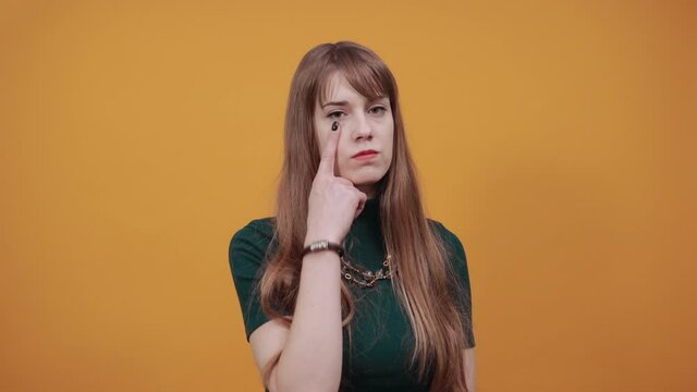 Contact lens on index finger, pointing eye with hand, thinking, having sensitive eyes, touching herself face by forefinger. Young attractive woman, dressed green shirt blonde hair, yellow background