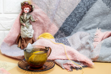 Green tea with lemon in a transparent dark mug on a yellow background and a knitted scarf with porcelain doll in english style. Cozy wallpapers, Home atmosphere, Copy Space
