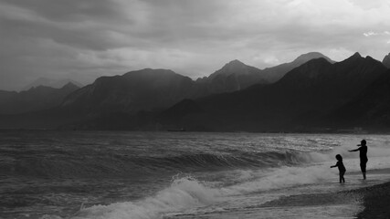 Silhouette of two kids enjoying the wavy sea on a rainy day in Antalya, Turkey. Black and white photograph of children, sea, mountains and clouds.