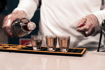 Un camarero joven llena unos vasos de chupito de una bebida alcohólica de color marrón en la barra de un bar mientras lleva guantes