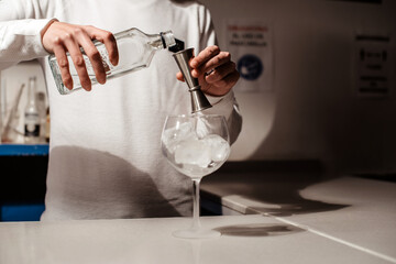 Un camarero joven vertiendo un alcohol blanco transparente en una copa con hielos sobre la barra de un bar usando un medidor