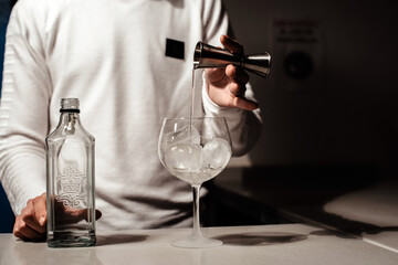 Un camarero joven vertiendo un alcohol blanco transparente en una copa con hielos sobre la barra de un bar usando un medidor
