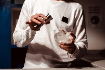 Un camarero joven vertiendo un alcohol blanco transparente en una copa con hielos sobre la barra de un bar usando un medidor