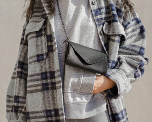 Woman wearing plaid trendy jacket on a gray hoodie with a black leather bag with her hand in her pocket.
