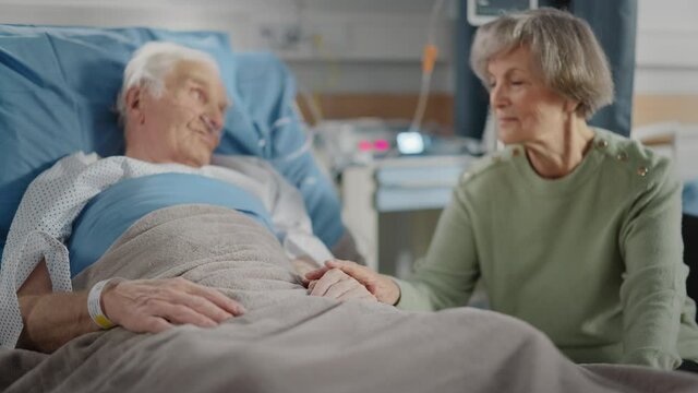 Hospital Ward: Elderly Man Resting in Bed, His Caring Beautiful Wife Visits, Supports Him Sitting Beside, Holding Hands, Happy Couple Talking Together