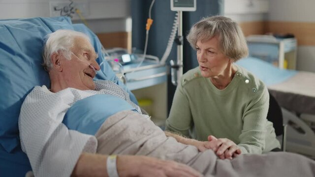 Hospital Ward: Elderly Man Resting in Bed, His Caring Beautiful Wife Visits, Supports Him Sitting Beside, Holding Hands, Happy Couple Talking Together. Old Man Recovering successfuly After Sickness