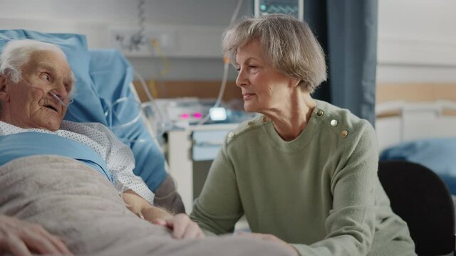 Hospital Ward: Elderly Man Resting in Bed, His Caring Beautiful Wife Visits, Supports Him Sitting Beside, Holding Hands, Happy Couple Talking Together. Old Man Recovering successfuly After Sickness