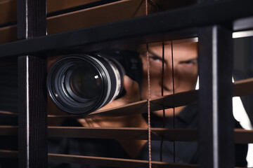 Private detective with camera spying near window indoors, focus on lens - obrazy, fototapety, plakaty