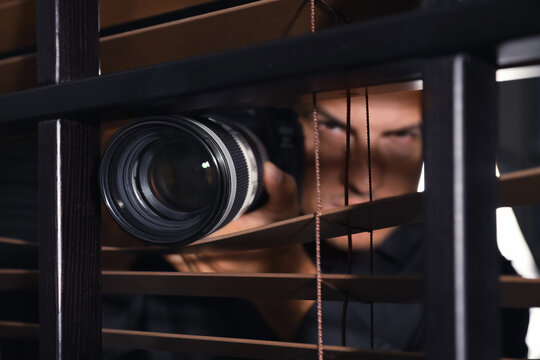 Private detective with camera spying near window indoors, focus on lens