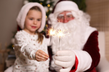 Santa Claus and little girl in room decorated for Christmas, focus on burning sparklers