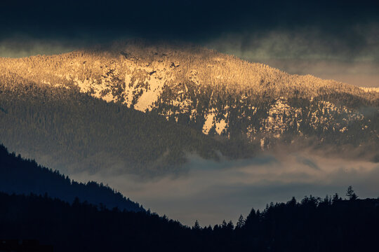 snow-covered mountain range with orange glow from the rising sun behind silhouette of the forest on the hill slope and thick layer of dark cloud in the morning 