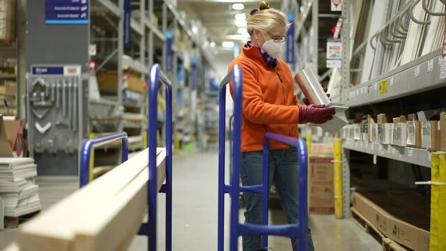 Mature woman wearing face mask looking at building materials in the lumber section of a big box hardware store and put them on a cart.