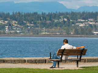 Rear view on a man sitting resting alone on wooden bench by the sea beach with mountain view. Nature relax outdoors.