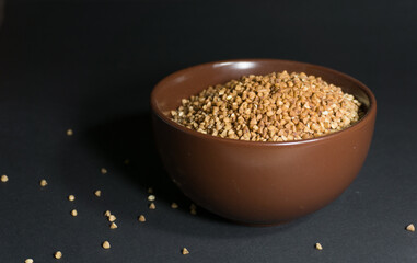 Brown uncooked buckwheat in a bowl with scattered grains nearby on a black background