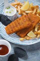 Close-up of breaded fish fillet with french fries, black potato chips and dipping sauces, vertical shot, selective focus