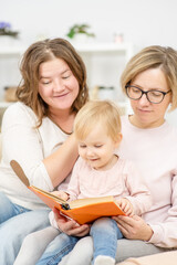 Obraz premium Two young women are sitting in the living room on the couch with a child on their knees and are reading a book. Gay lesbian couple at home with a baby.