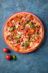 Pizza with bacon, spicy pepperoni, mozzarella, cherry tomatoes, mushrooms, red onion and sweet pepper on blue background. Copy space, vertical composition, top view