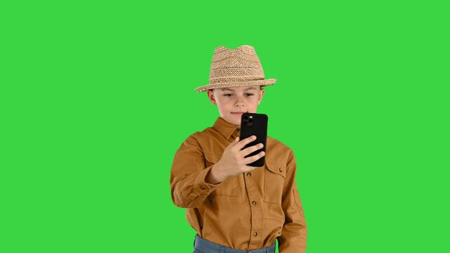 Little boy in a straw hat taking selfies on his phone on a Green Screen, Chroma Key.