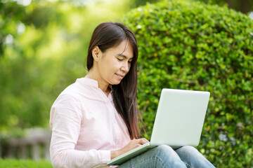 Asian woman sitting green park using laptop computer. Woman working on laptop happy entrepreneur business using notebook with hands typing on keyboard home office during coronavirus quarantine period