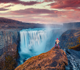Photographer takes picture on the cliff on falling water of most powerful waterfall in Europe - Dettifoss. Breathtaking summer sunrise in Jokulsargljufur National Park, Iceland.