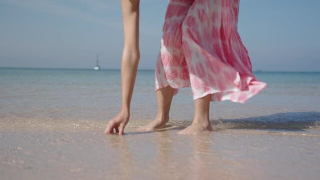 Shot of Female bending down picking up clam shell on white sand beach on sunny day. Young pretty girl playing with sea sand water and waves. summer vacation. Low camera angle, slow motion wind blowing
