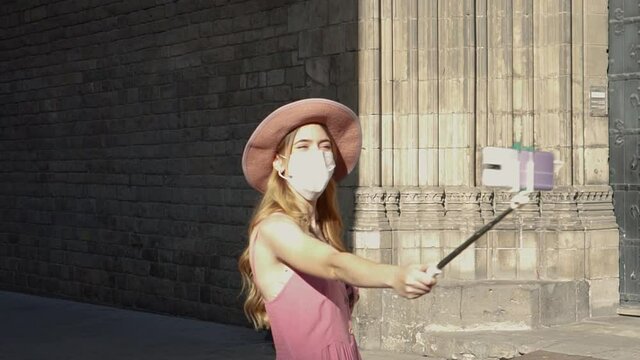 Young tourist woman taking a selfie with her phone wearing a face mask due to coronavirus pandemic. Girl using her smartphone to take pictures of herself walking in the city of Barcelona 
