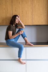 Attractive woman sitting on kitchen counter holding glass of juice. Young housewife relaxing in the kitchen.