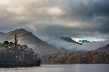 Fototapeta premium Dramatic landscape image looking across Derwentwater in Lake District towards Catbells snowcapped mountain with thick fog rolling through valley