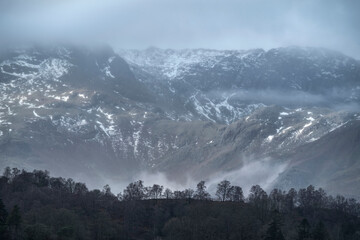Epic dramatic landscape image of view from Elterwater across towards Langdale Pikes mountain range on foggy Winter morning