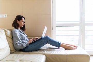 Side view of casual young woman using laptop on sofa at home. Working from home in quarantine lockdown. Social distancing Self Isolation