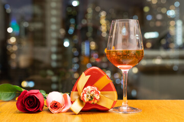 glass of red wine, pink rose, heart-shape red candy container on wood table with window view of city buildings in background