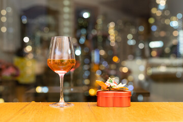 glass of red wine, heart-shape red candy container on wood table with window view of city buildings in background.