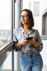 Fototapeta premium Excited beautiful woman in eyeglasses using smartphone while sitting near window