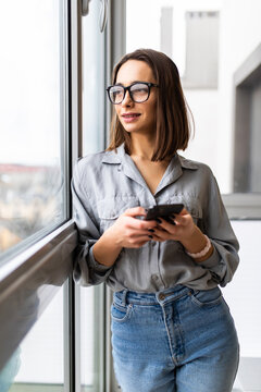 Excited beautiful woman in eyeglasses using smartphone while sitting near window