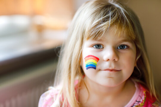 Portrait of cute little toddler girl with painted rainbow with colorful colors on face. Lonely child during pandemic coronavirus quarantine. Children make and paint rainbows around the world