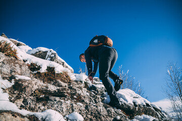 Young mountaineer walks and climbs on the snowy rocks of a mountain with backpack on his shoulder during a sunny winter day